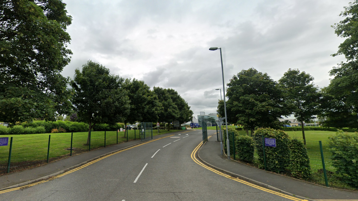 A road leading to a Haughton Academy, a glass building can be seen surrounded by grass and trees.