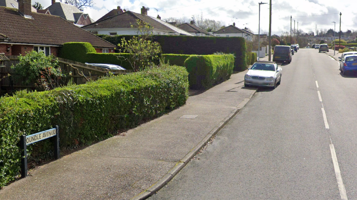 A streetview of Oundle Avenue. Houses along the street have green shrubs lining the pavement. There are cars half parked along the pavement too.