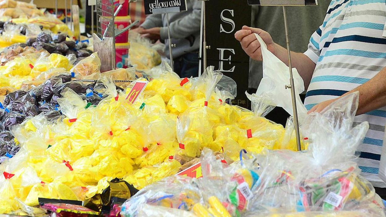 Man serving in front of a stall filled with yellowman in bags, alongside other sweets