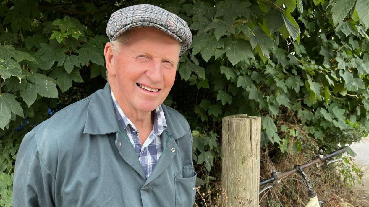 An older man in green overalls smiling at the camera. He is standing in front of a green bush.