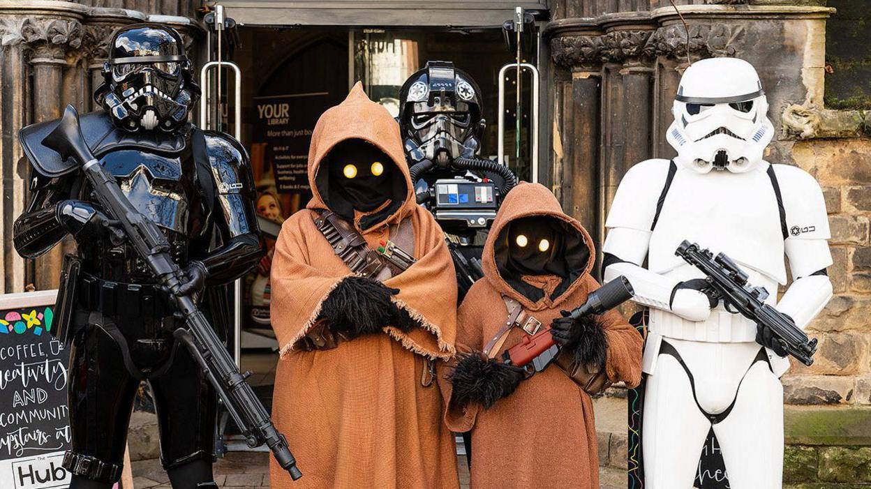 Three people dressed as Stormtroopers and two dressed as Jawas from Star Wars, pose for a picture outside of the entrance to a building.