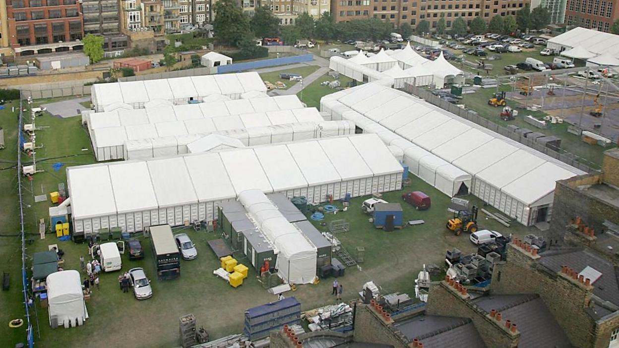 Aerial view of the temporary mortuary, featuring numerous long white tents arranged in rows on a grassy field. The site is surrounded by vehicles and equipment, such as bright yellow bins.
