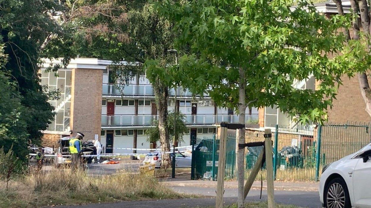 An area in front of a small block of flats is cordoned off with blue and white police tape. There is a police officer in a black hat and fluorescent yellow vest guarding the tape. A forensic officer in a full white suit is looking inside a black car