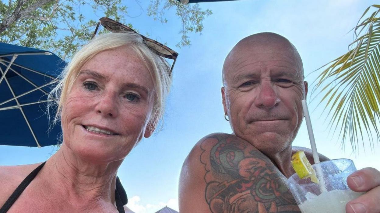 Head and shoulder shot - selfie - of a middle-aged couple. The woman, on the left, has tied up blonde hair and sunglasses pushed up on top of her head. The man is bald with a large shoulder tattoo. He is holding a glass containing what appears to be a cocktail with a straw. Both are smiling and it is in a holiday setting, with blue sky behind them and parts of a sun umbrella and a palm visible.