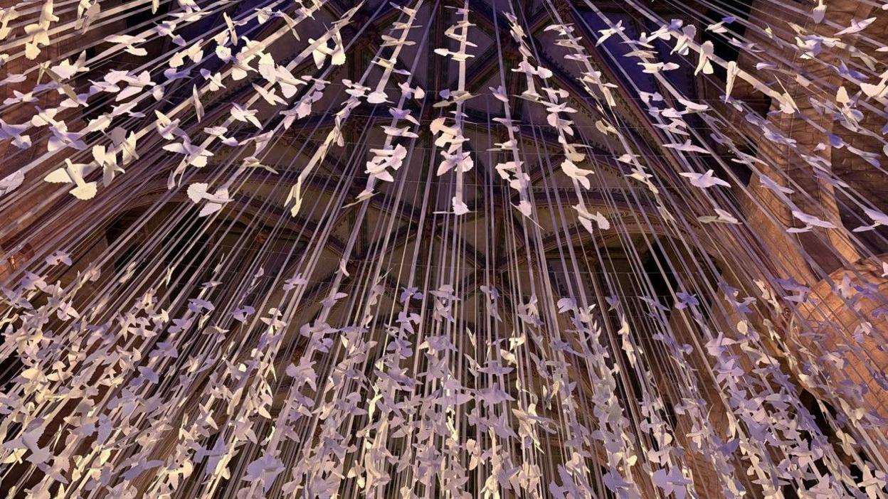 A close-up image of thousands of paper doves hung from the ceiling of an abbey with purple lighting shining over them.