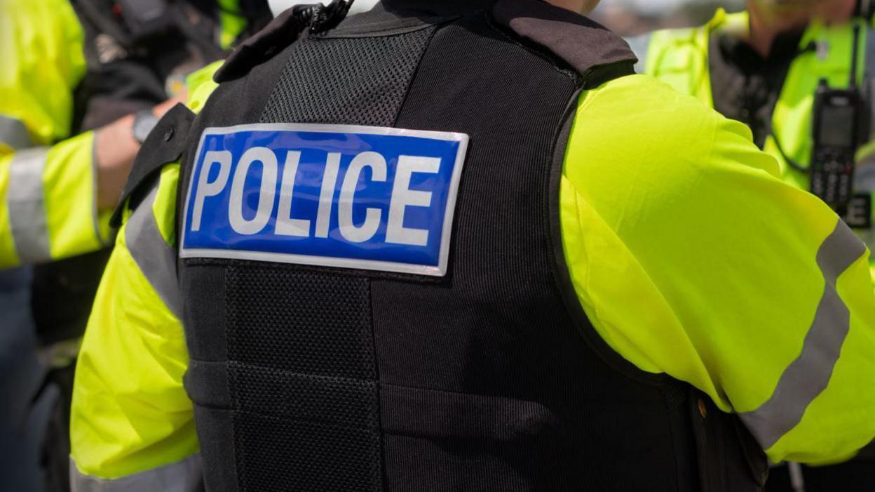 The back of a person in a black stab vest with the word police on the back, with a yellow reflective jacket underneath