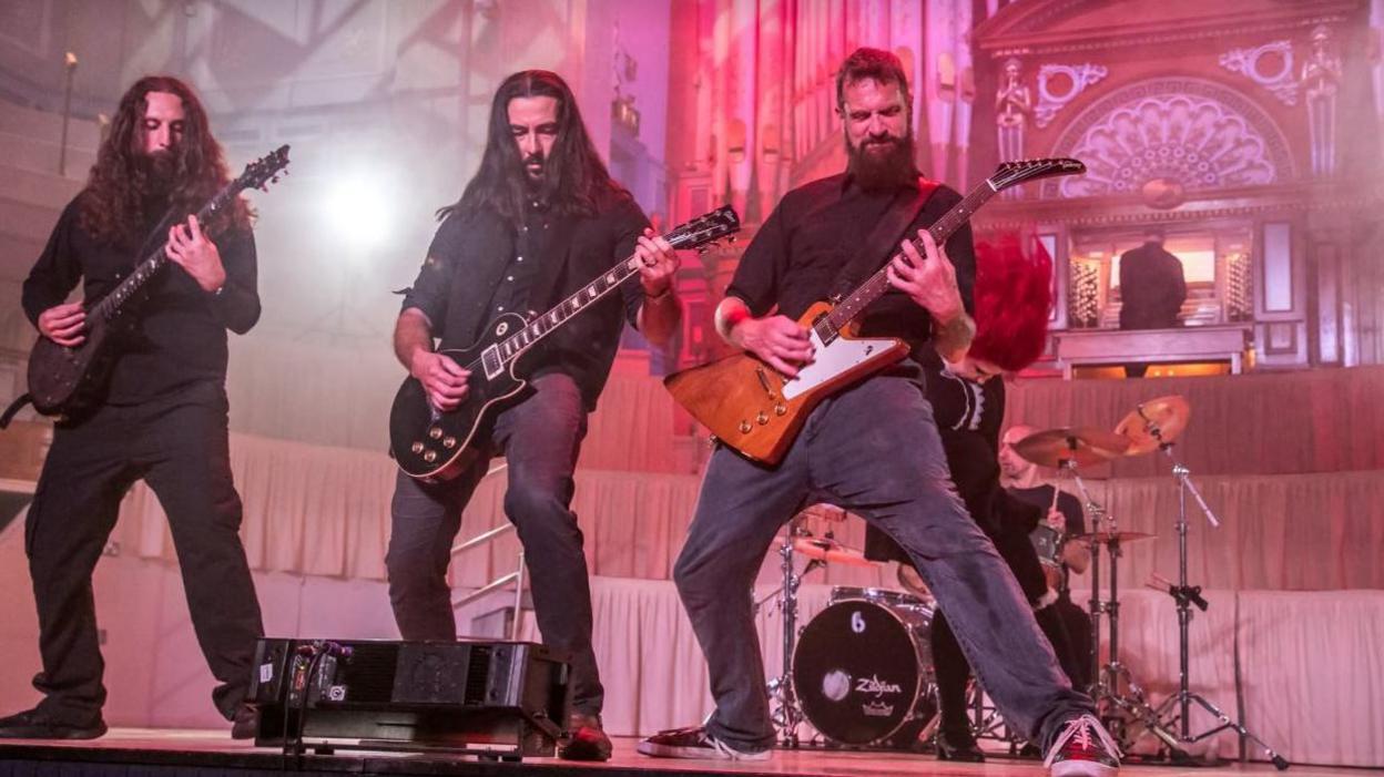 Two long haired men with guitars and one short haired man with a beard play guitars in front of a red haired wearing a black blouse with a white collar. In the background a man plays a church organ.