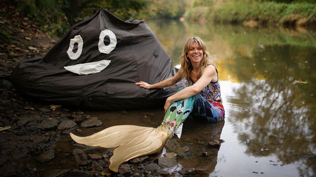 Woman in a mermaid costume sitting on rocks in a shallow part of a river with a big brown beanbag looking like the poo emoji next to her