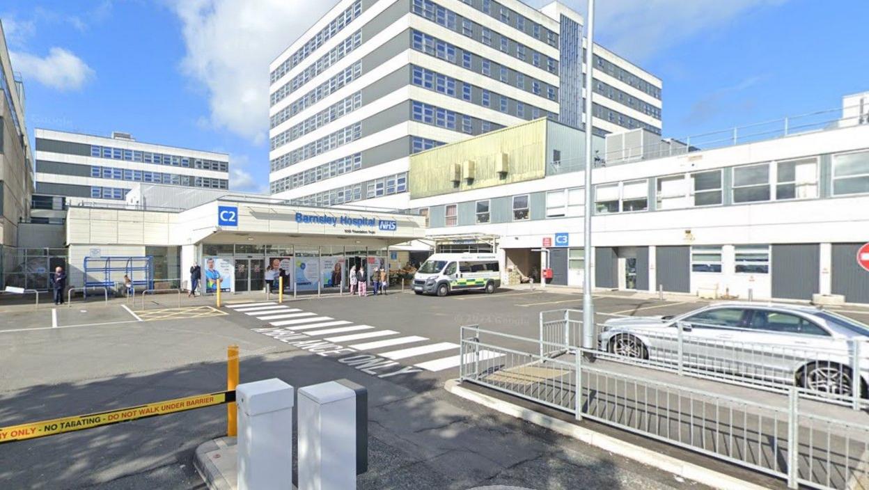 The outside of the front entrance to Barnsley Hospital. The entrance is a single storey building with people milling about outside, while an ambulance sits in close proximity. Hospital buildings of varying heights can be seen dotted around in the background.