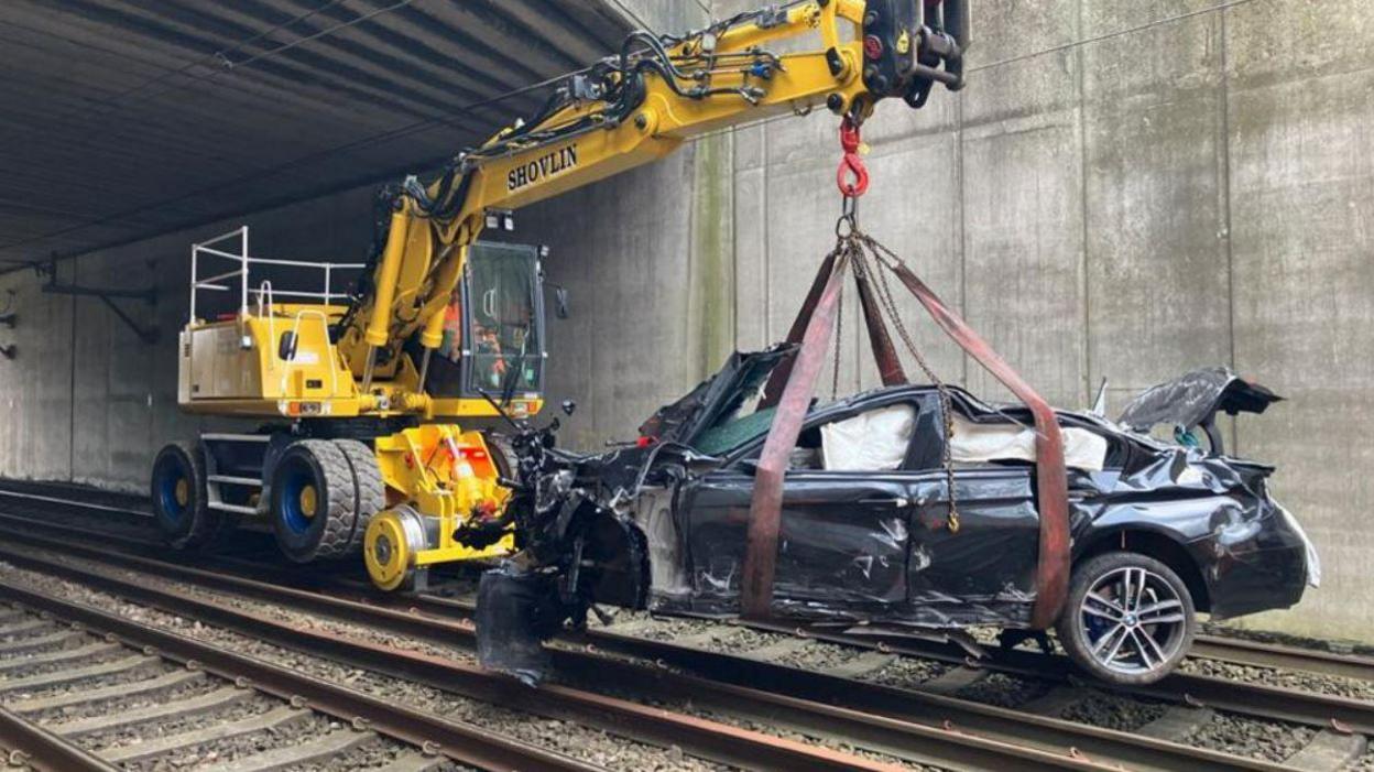 A yellow cherry-picker winches a smashed black BMW off the tracks