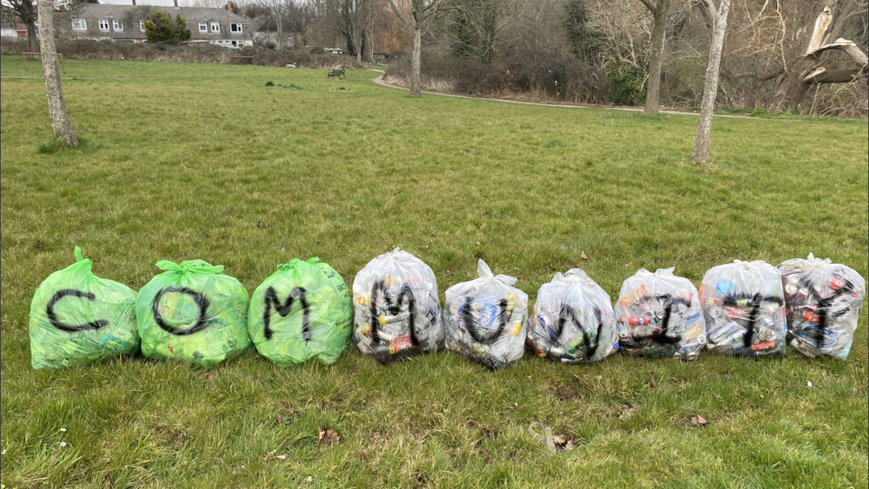 A row of nine full litter bags sitting on the grass in a park with the word 'community' spray-painted across them in black paint.