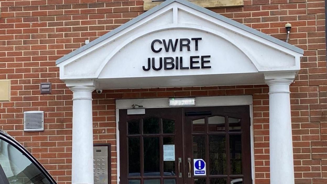 The outside of the Cwrt Jubilee building with a car parked on the left. There are brown doors which have a warning sign for the automatic doors and there are white pillars and across the front in black script it says "Cwrt Jubilee"