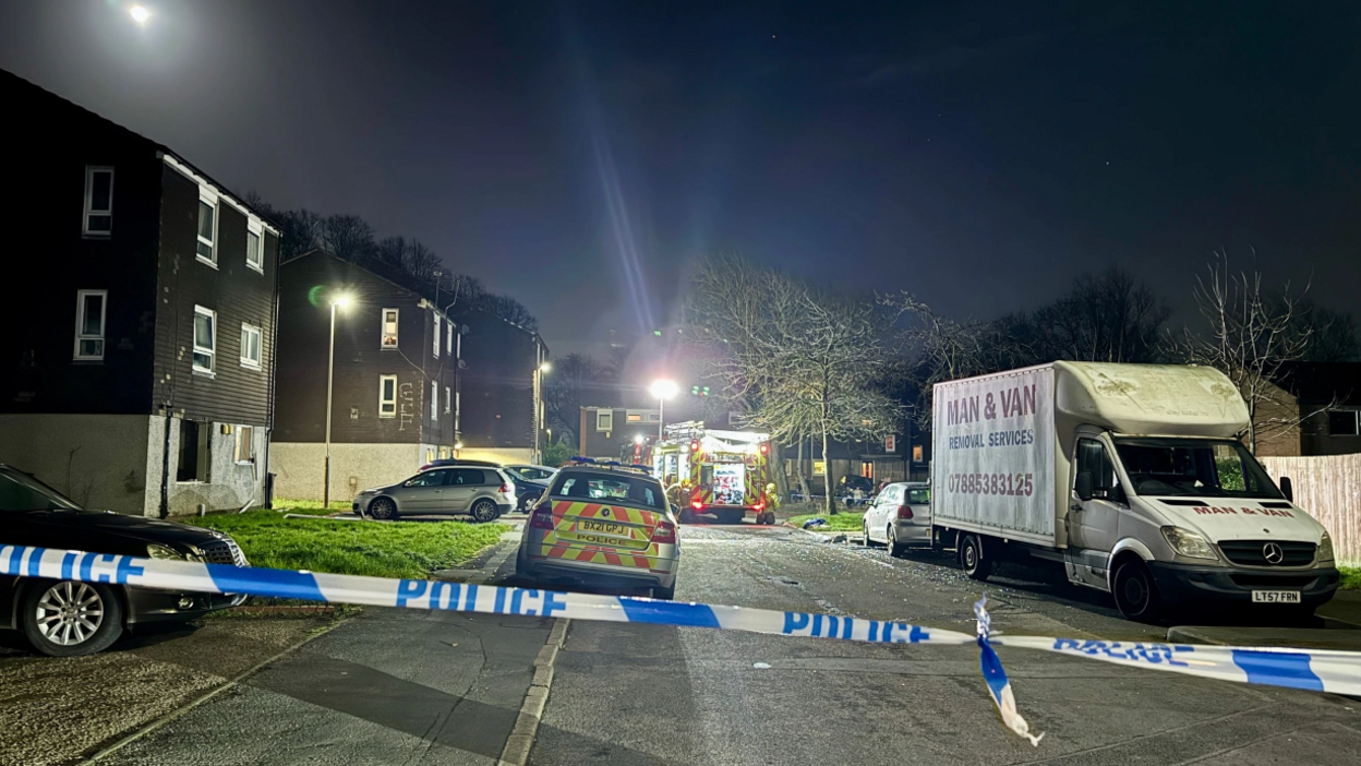 Police and fire service vehicles inside a police cordon in a Leicester street, taken at night. A flat on the left of the images is windowless with shattered glass on the road in front of it.