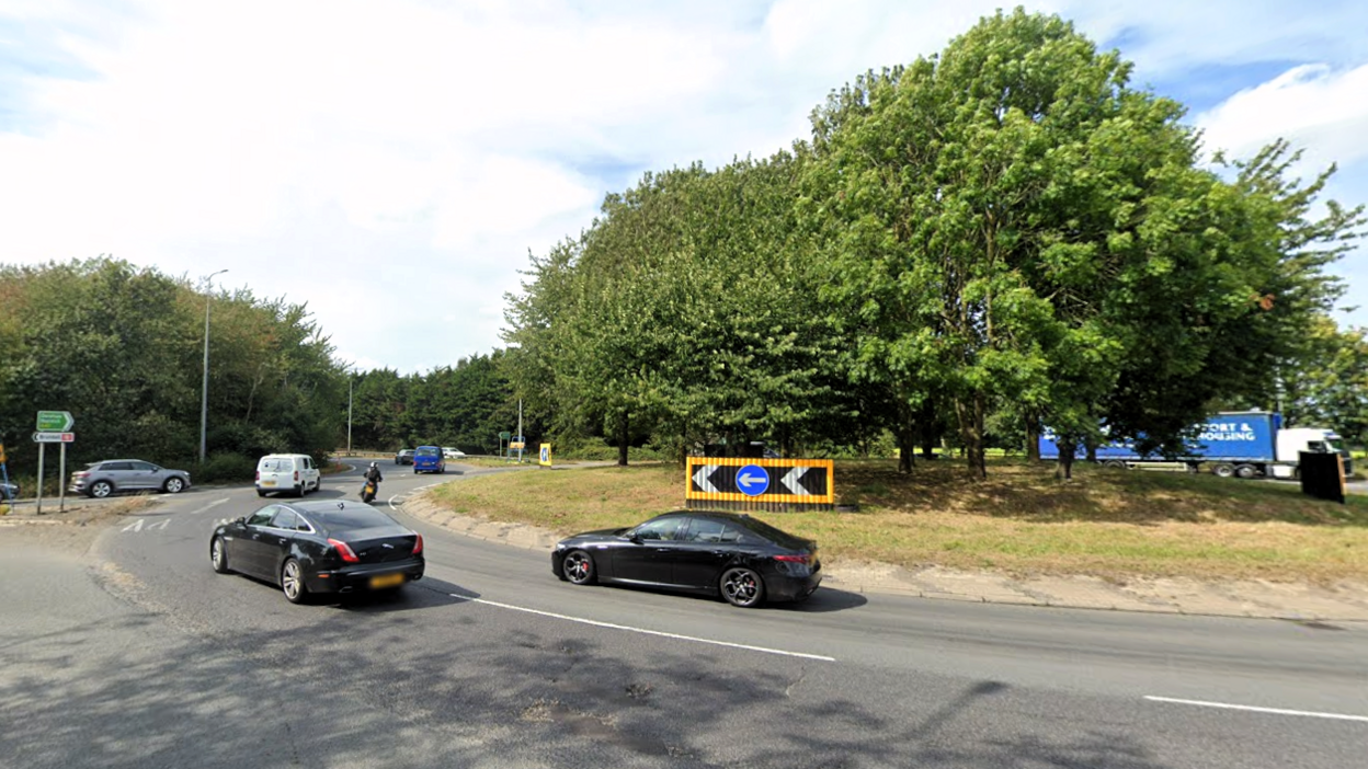 A roundabout with two black saloon cars circling it. A white van, motorcycle and other traffic can be seen in the distance. The middle of the roundabout is covered in grass and trees.