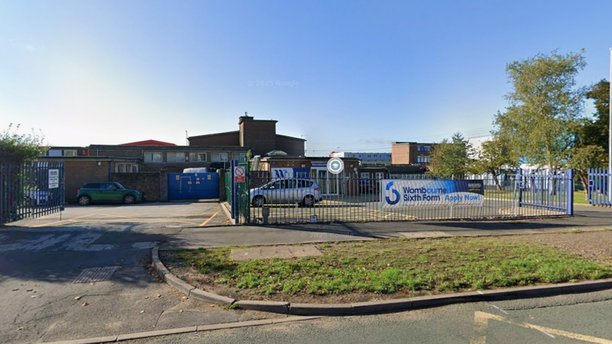 Bright blue sky can be seen with mainly brick, school buildings in the lower half of the image. A car park is surrounded by a fence which stands on the pavement with a grass verge on the other side. A banner advert for sixth form places is tied to the fence.
