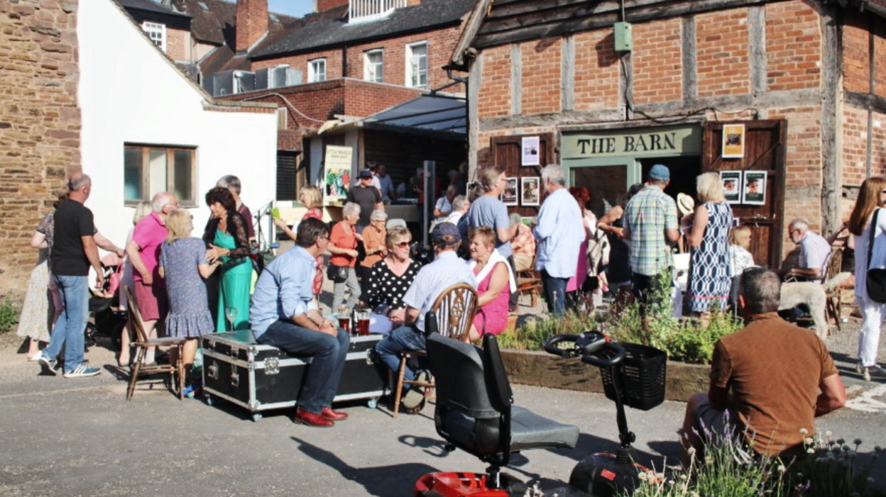 A number of people, some stood, some sat, outside a brick and timber building in the sunshine. The building's doors are open, and a sign above them reads The Barn