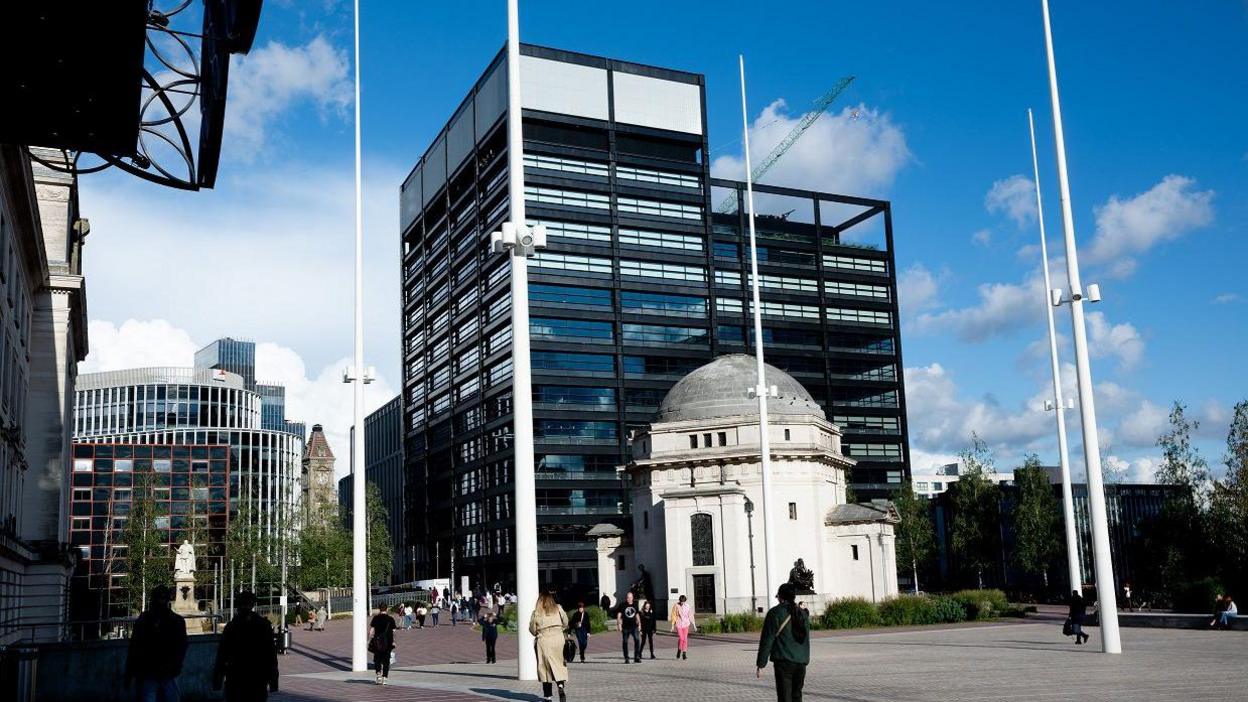 A large glass office building that is about 14 storeys tall. Its exterior is black and white, and there is a roof garden on its back right hand side. In front of it, is a smaller, white domed building.