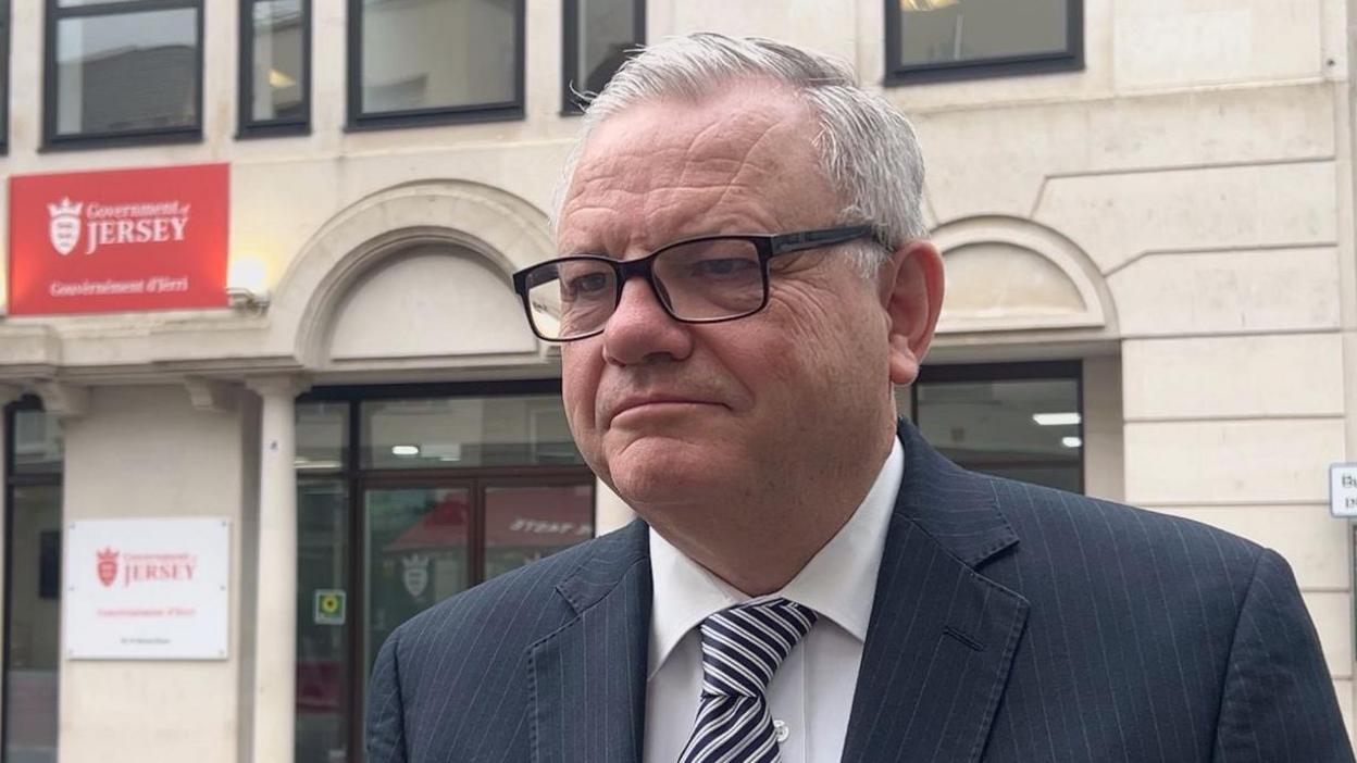 Constable Richard Vibert is wearing a black pin stripe suit with a white shirt. He is wearing glasses and is stood outside the Government of Jersey building on Broad Street. 