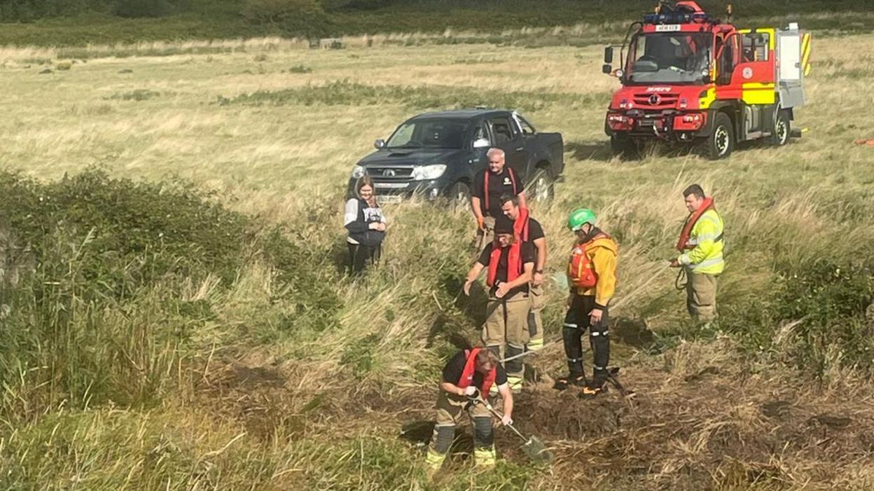 Firefighters, a car and a specialist fire service all-terrain vehicle on farmland in Suffolk. One of them is using a spade to dig away at the riverbank.
