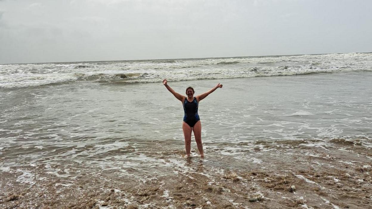 Helen Jewell in her swimming costume, standing in the shallows of the sea with waves behind her