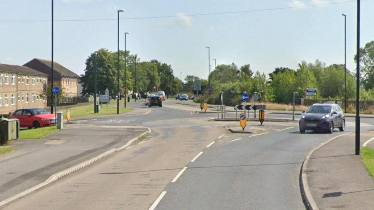 A road leading to a roundabout. Cars in the distance, trees on either side of the road. Houses on the left hand side of the road.