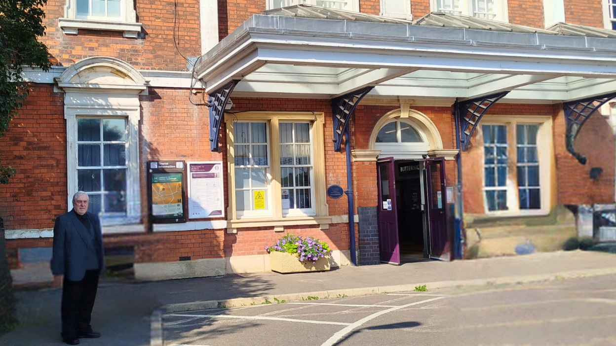 Phil Knowles in a suit standing to the side of the front entrance to a red brick railway station with a purple door and an ornate porch