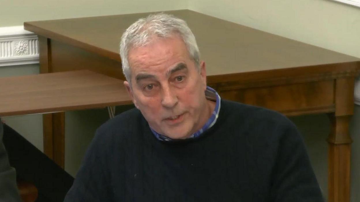 Pat Sheehan is wearing a blue shirt and navy jumper. He has grey hair. In the background there is two wooden tables. The walls are painted green and have a white feature running along them.