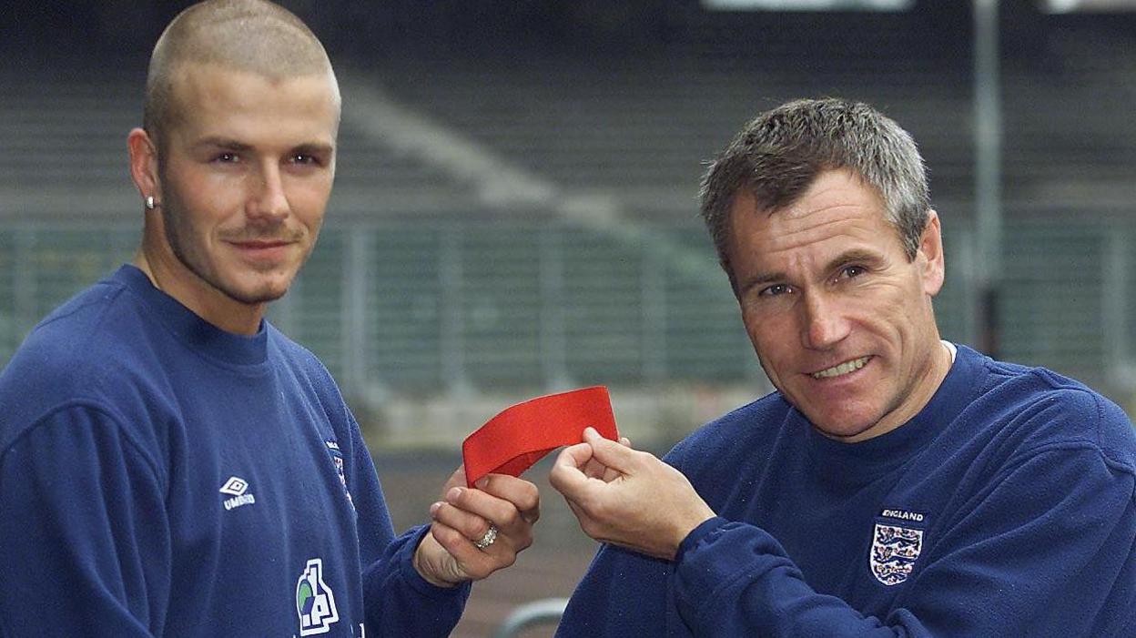 An earring-wearing, shaven-headed David Beckham is handed a red England's captain's armband by grey-haired coach Peter Taylor in 2000, with both men wearing England tracksuits