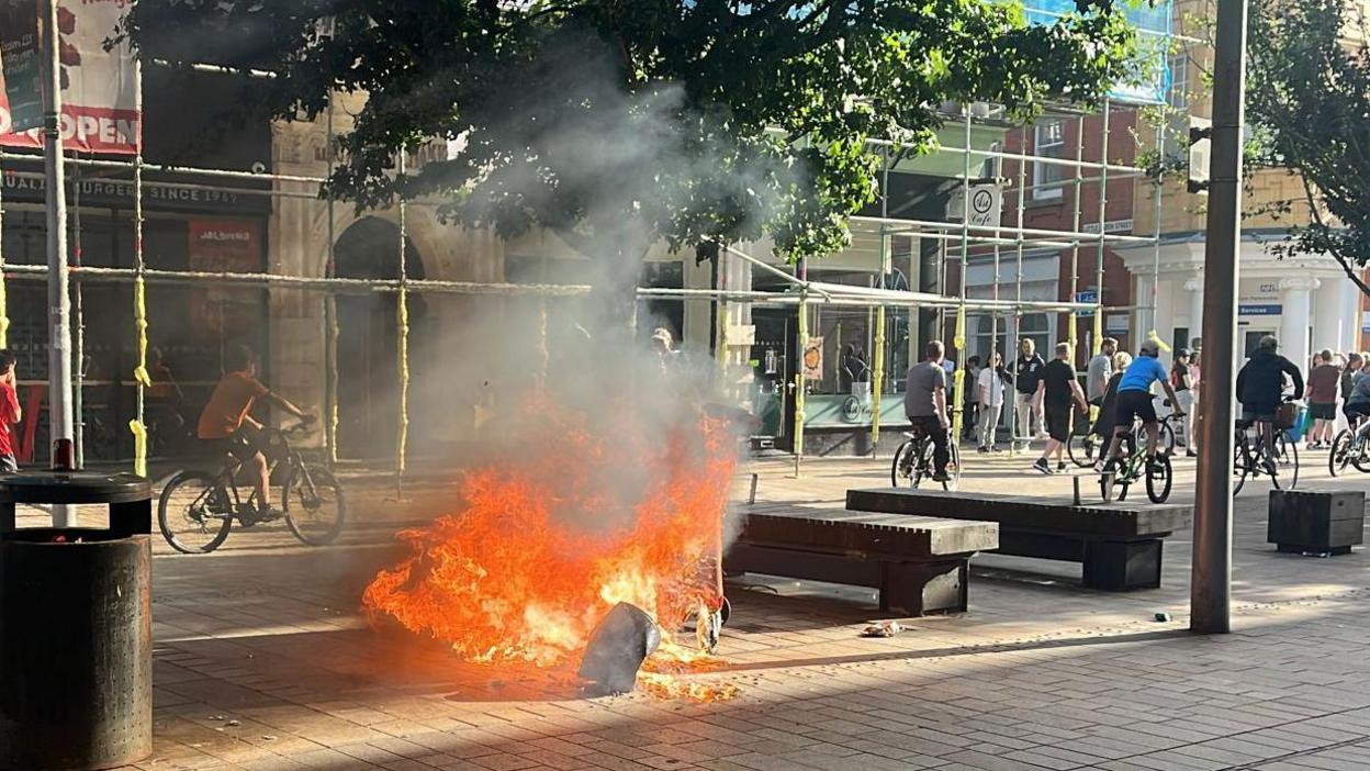 Bin on fire in the city centre with people on bikes cycling away