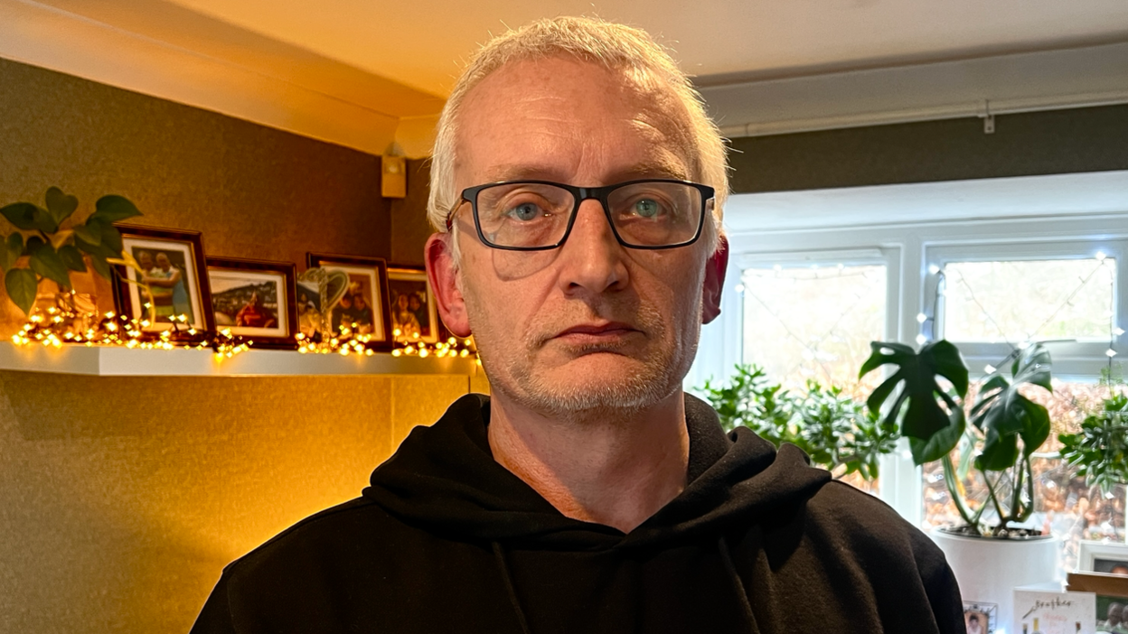 John Franklin with cropped white hair and glasses. He wears a black hoodie and stands inside a room with photographs and plants on shelves