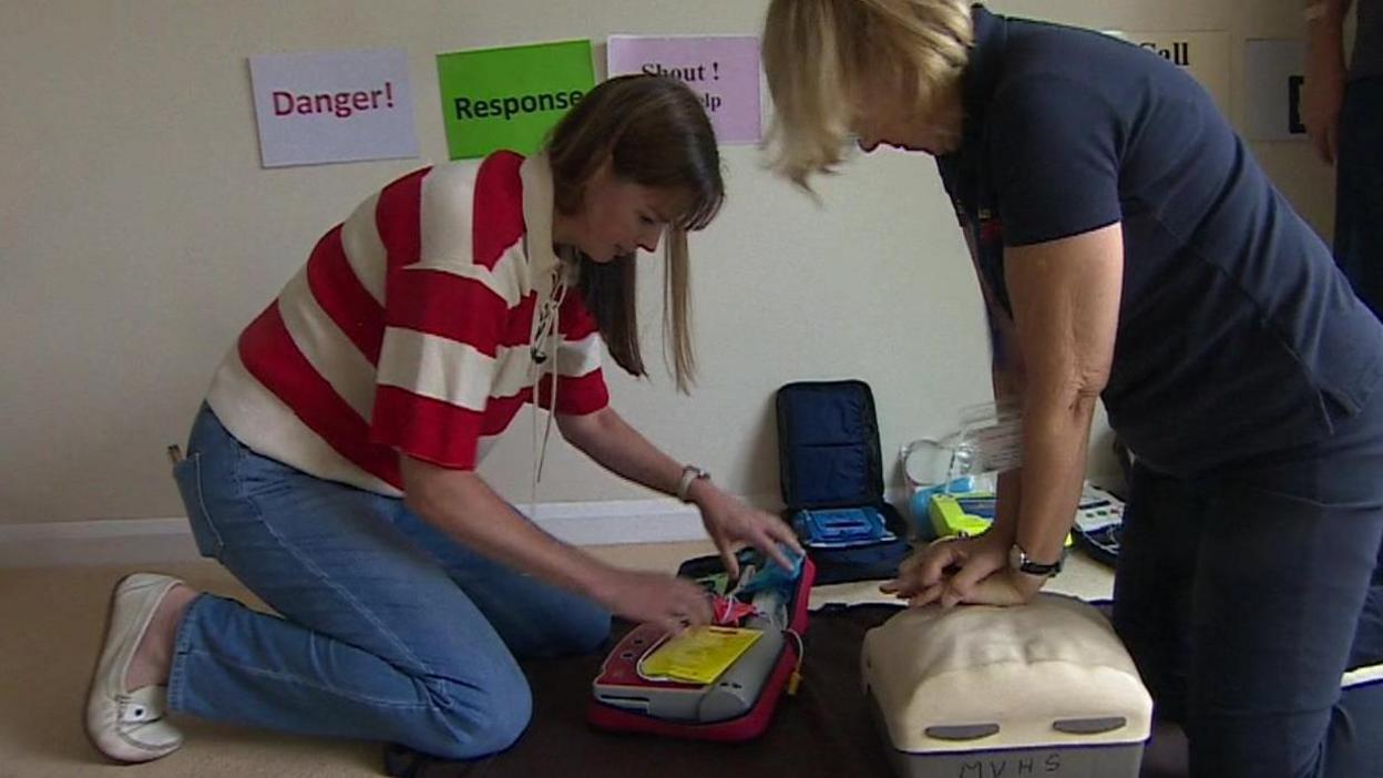 Kneeling, Alexis Green works on a defibrillator unit while another woman performs CPR on a medical dummy.