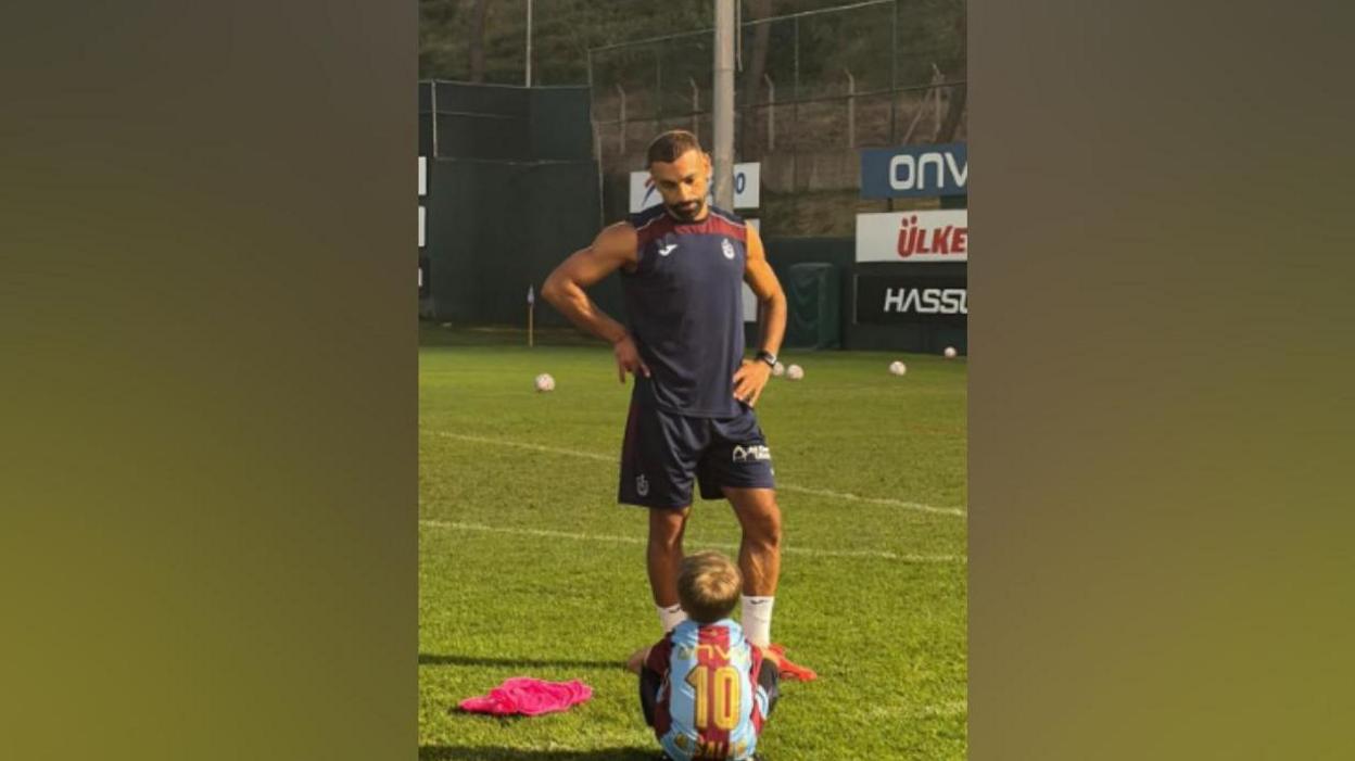 Image shows Isaac sitting on a training football pitch. Salah stands in front of him with his hands on his hips. 