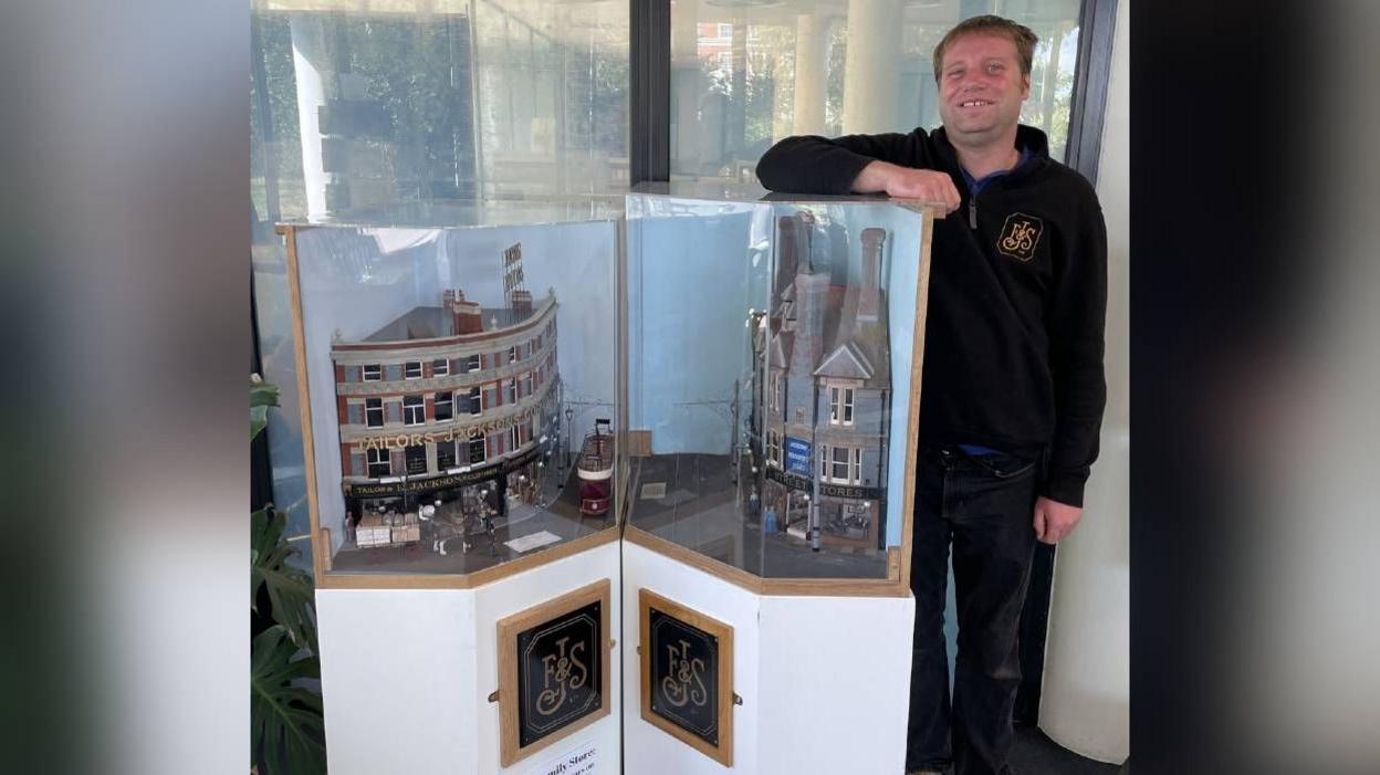 Thomas Macey stood behind a cabinet which has two models of the stores inside. The intricate and realistic models of buildings are as follows: The one on the left has a curved redbrick facade and says "Jacksons Corner" on it, with a tram and a horse and cart ouside. The one on the right says "Duke Street stores" on it and is made of grey brick.