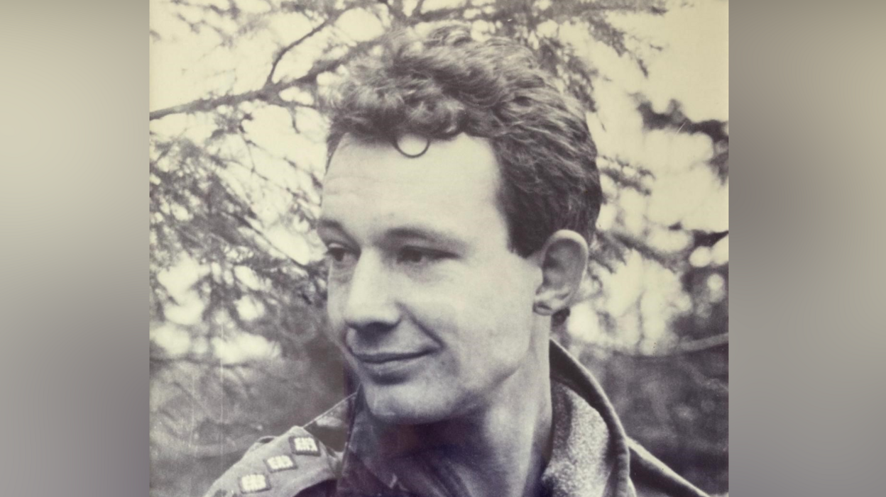 A black and white picture of Major Christopher Dockerty. He is smiling as he looks away from the camera. He has short hair and a shaved beard. He wears an army uniform.