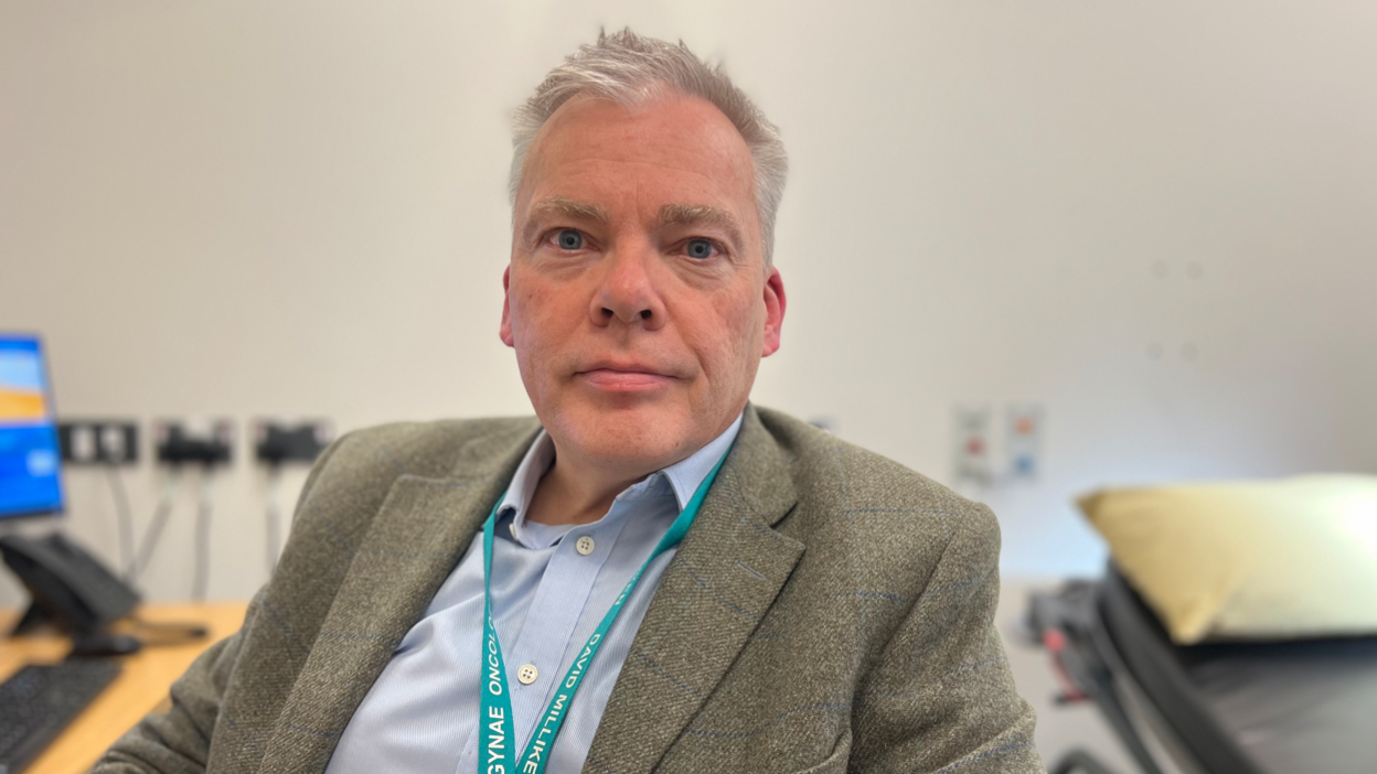 A close-up of David Milliken in a clinical setting wearing a smart jacket, blue open-necked shirt and turquoise lanyard. There is a computer and a patient bed behind him