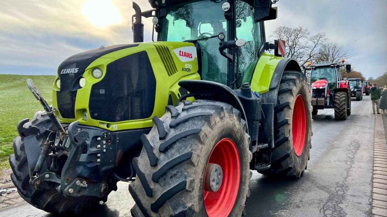 A brightly coloured tractor leading a procession of tractors on a rural road near Beverley as supporters line the route.