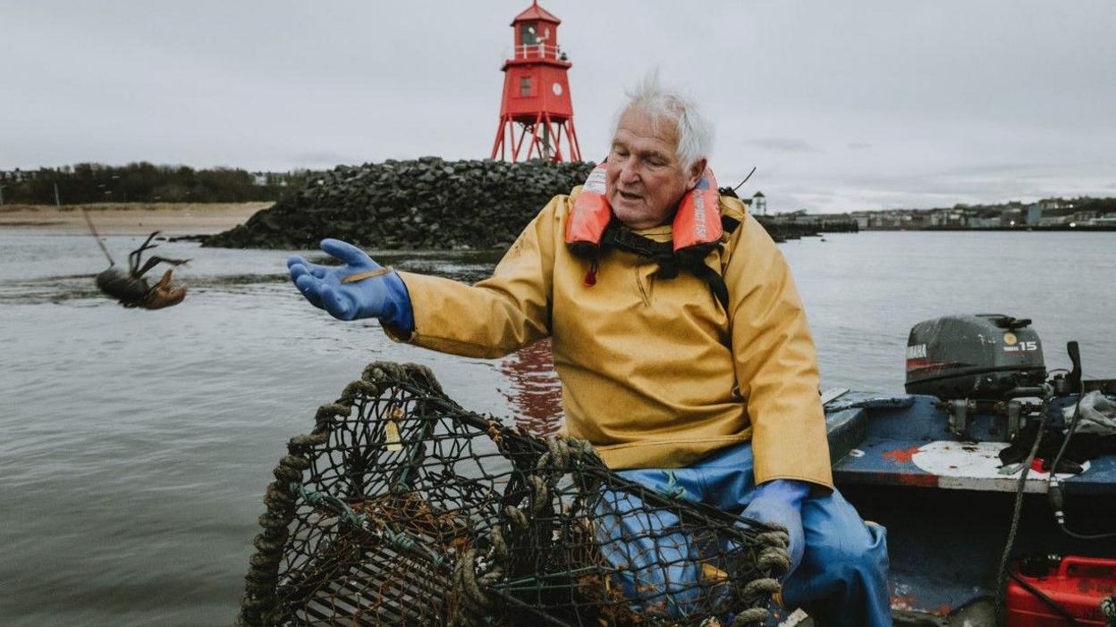 A white-haired man in yellow water proof clothing is sitting on a boat and throwing a lobster back into the sea. He is not far from the shore.