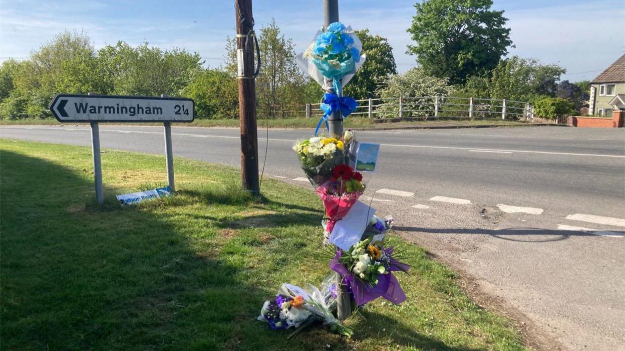 Flowers and cards tied to a lampost by a road junction in a rural area. A road sign for Warmingham can be seen on a verge at the site. 
