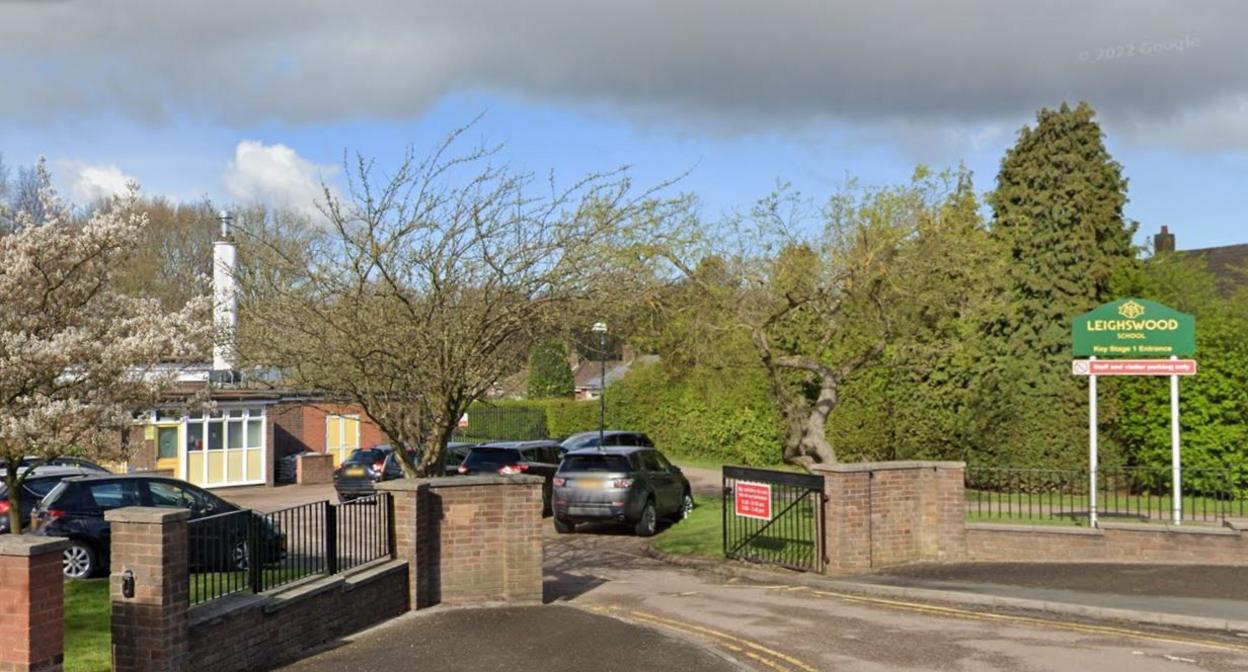 A single-storey building, with a small car park and a fence at the front of the building. The building has yellow doors and window frames. There are black and grey cars in the car park. There is also a green sign near the car park that says 'Leighwood School' in yellow.