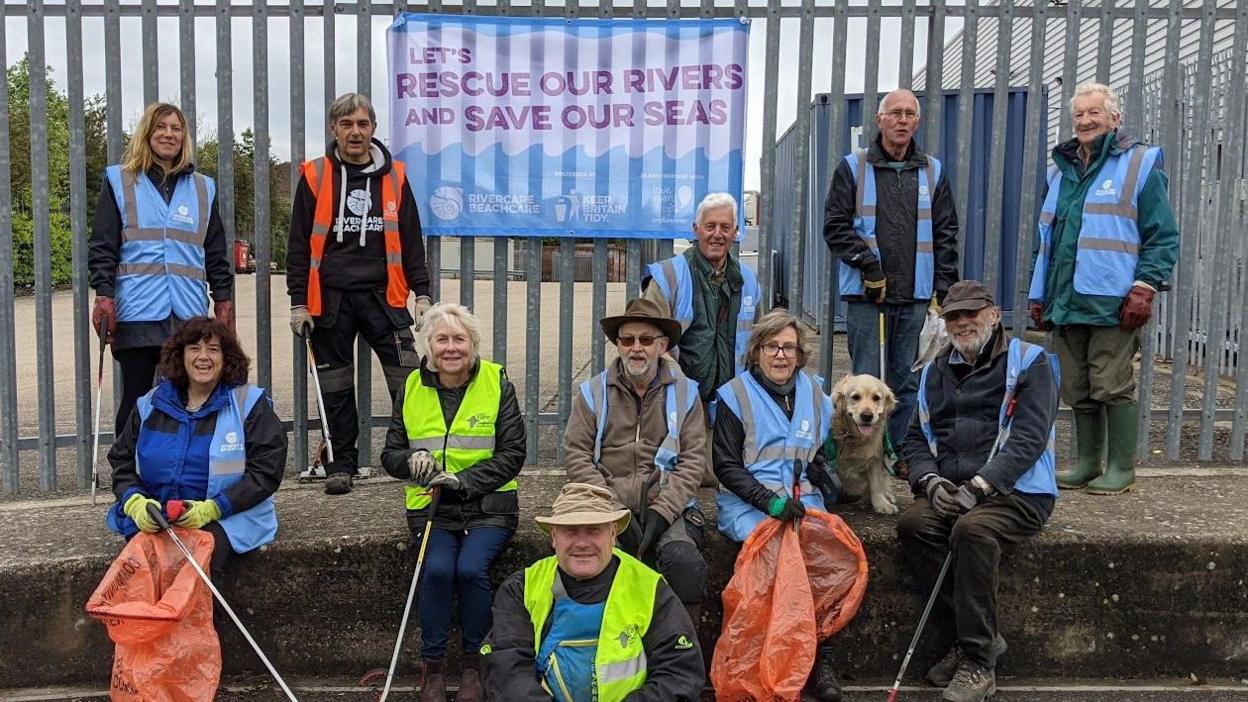 A group of litter pickers wearing blue, yellow and orange high vis vests, with some standing on top of a wall and others sitting below it. They are holding litter picks and bags and there is also a golden retriever. A sign on the fence says 'let's rescue our rivers and save our seas'.