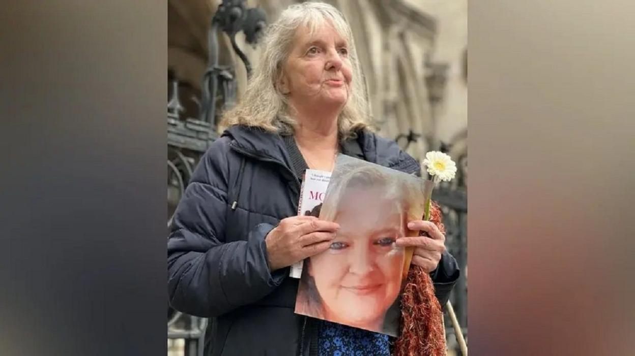 Joy Dove stands outside the Court of Appeal in London. She has shoulder-length grey hair and is wearing a blue coat and is holding a photograph of her daughter Jodey Whiting and a yellow and white flower