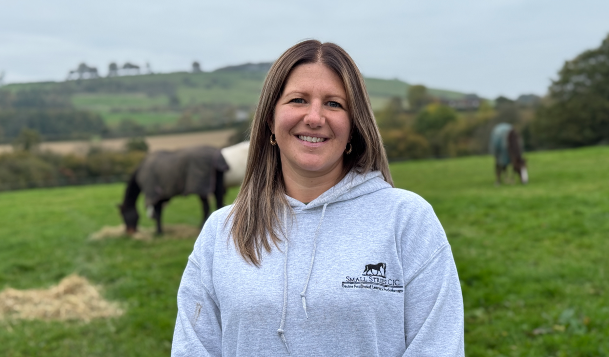 A woman with mid-length brown hair in a grey sweatshirt stands in front of three horses grazing.