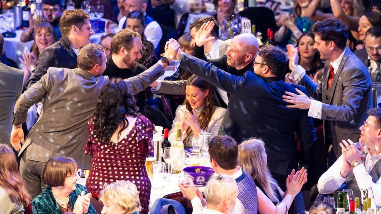 Members of the band celebrate with friends and family. They are high fiving each other while other guests look on smiling.