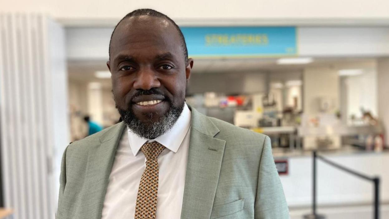 Marvin Charles looks into the camera. He is stood in the canteen at his school, in front of the large serving hatch where his pupils have just been in for lunch. He is smiling and wearing a light green suit over a white shirt, with a yellow and brown tie.