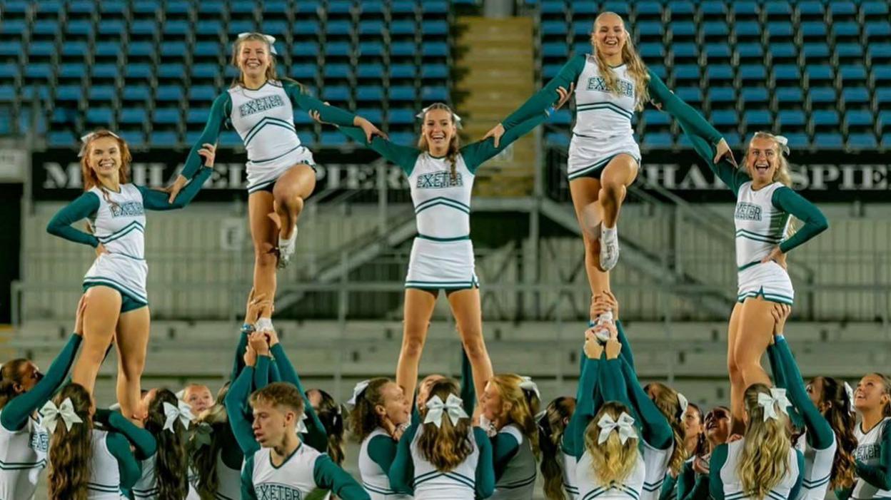 Cheerleaders stand on the shoulders of their team mates in formation. They are wearing green and white uniforms.