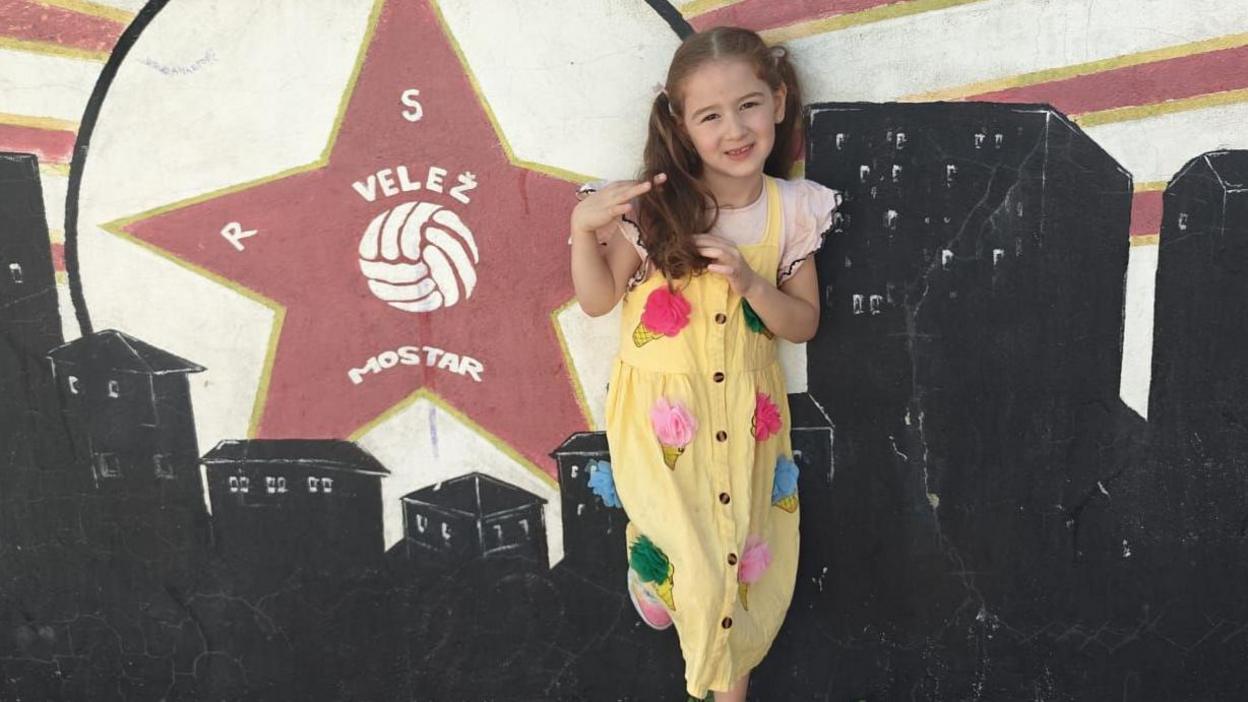 A child is standing outside against a mural painted on a wall. The mural features a large red star with the text “VELEŽ MOSTAR” and a volleyball graphic inside it, along with stylized city buildings in black and red rays radiating from the star. The child is wearing a yellow dress decorated with colorful circular patches and white sneakers with pink details.