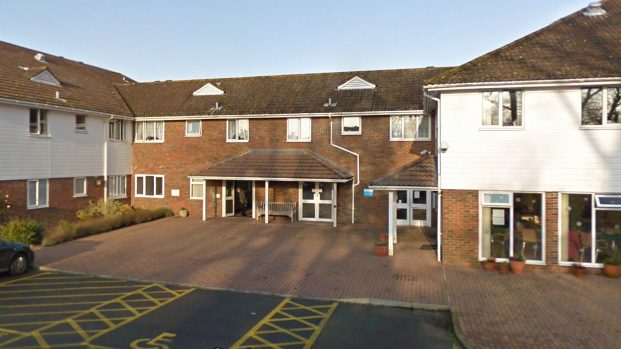 The entrance to Ticehurst Surgery, a collection of brown brick two-storey buildings with white cladding to the left and right and spaces for disabled parking opposite the main door. An elderly man can be seen leaving the building