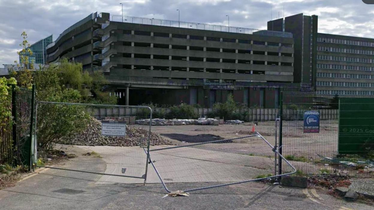 The Greyfriars site in Northampton, with metal gates across the entrance. A concrete surface is visible behind the gates, and a multi-storey car parking in brick and concrete is beyond. The sky is cloudy and grey.