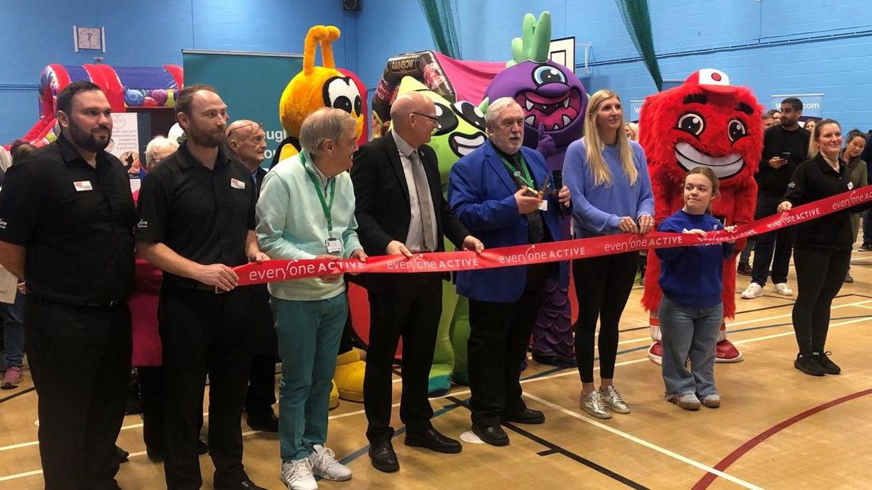 A line up of council and leisure centre representatives, alongside two celebrity athletes and a group of weird looking mascots, cutting a giant ribbon in a sports hall