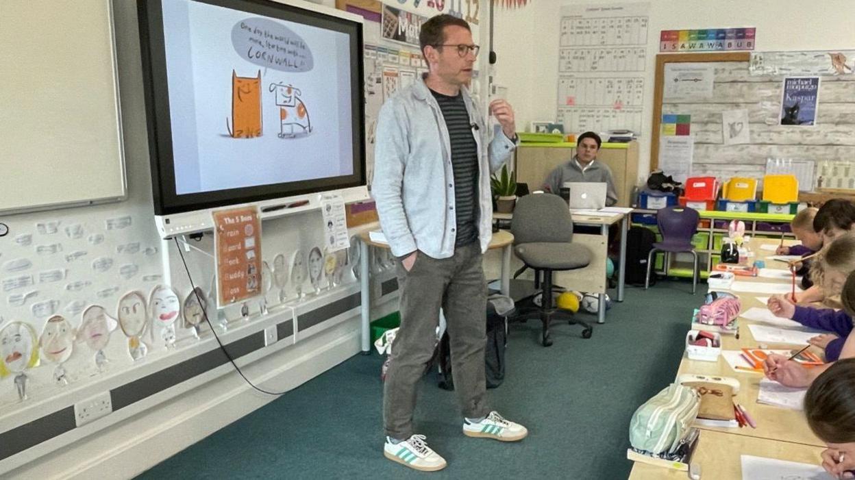 Author and illustrator Tom McLaughlin stands in front of a smart board with a sketch of a dog and an orange cat on it. The children sit at desks in front of him, learning to draw their own animals, for their own comic books that they are making.